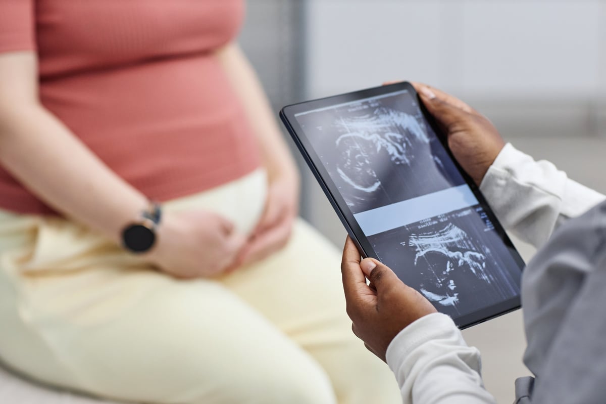 Black woman holding digital tablet displaying ultrasound images while sitting near pregnant Caucasian woman with hands resting on belly during prenatal medical consultation