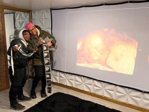 Two people in winter coats standing on a ladder in front of a projection screen displaying warm-toned imagery